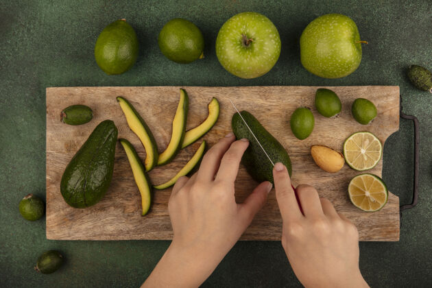 视图俯视图中 女性用刀在木质厨房板上切割成熟新鲜的鳄梨 绿色墙壁上隔离着酸橙和青苹果肥肉新鲜苹果