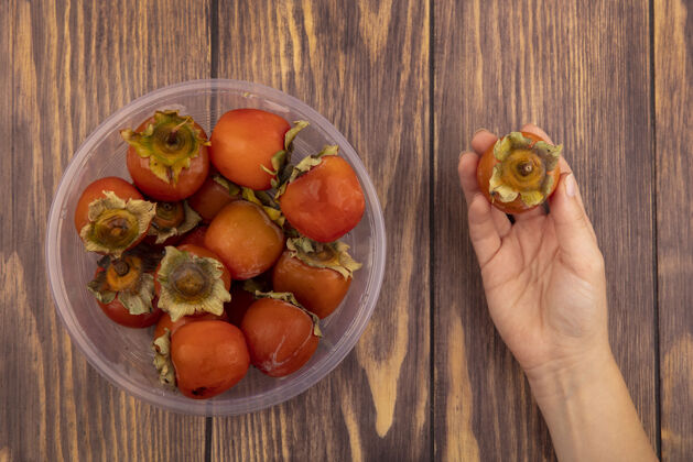 明确俯视图：女性手拿着一个成熟柔软多汁的柿子 柿子放在一个透明的塑料碗里 放在木墙上木头柿子新鲜