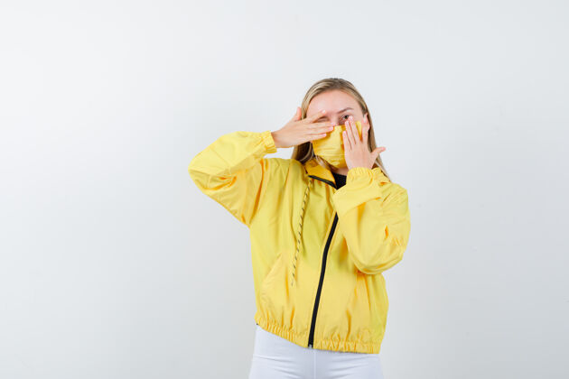 健康照片中的年轻女士手放在一只眼睛上 穿着夹克 裤子 戴着面具 看着好奇的前景色眼睛护理夹克