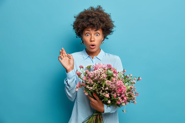 奇迹震惊的卷发非裔美国女士收到未知的人的花束 盯着的眼睛 因为得到意外的交付 穿着时尚的蓝色衬衫 站在室内花卉概念花姿势问候