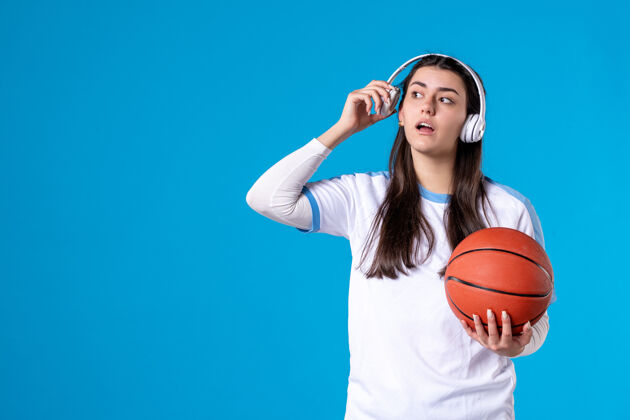 球员正面图：戴着耳机拿着篮球的年轻女性站在蓝色的墙上篮球球前面