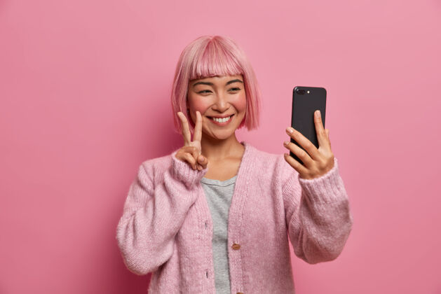 快乐微笑真诚的亚洲女人做出和平姿态 在视频会议中欢迎朋友 通过智能手机自拍 穿着休闲套头衫 留着时髦的染发 毛衣女孩和平