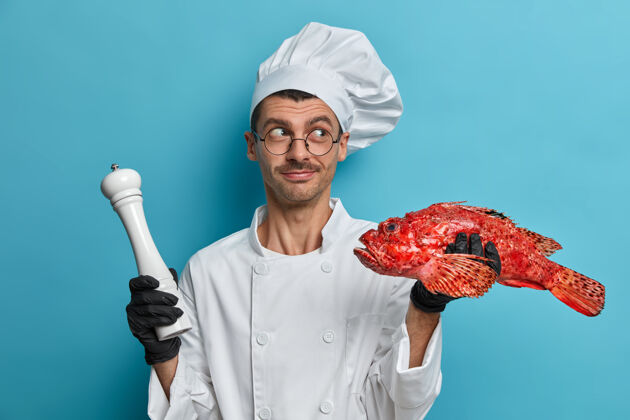 鲈鱼图为专业男厨师手持生红海鲈鱼和胡椒粉磨调味 身穿厨师制服晚餐人鱼
