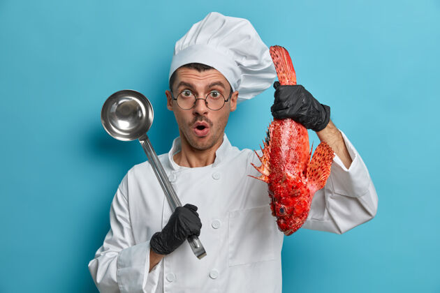 鱼图为惊讶的男厨师尝试最好的鱼食谱 手持红海鲈鱼 勺子 戴着厨师帽和白色制服红色室内人