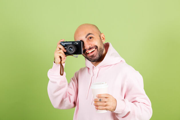 手势年轻秃顶的白种人 穿着粉色连帽衫 正拿着相机和咖啡享受男人纸杯
