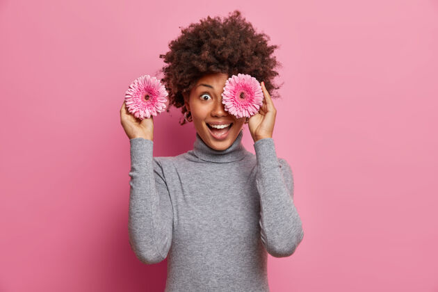 微笑快乐的非裔美国女人用玫瑰色的非洲菊花遮住眼睛 开心地咯咯笑着 准备去装饰派对的房间雏菊华丽发型