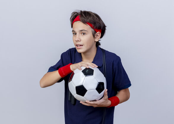 周围令人印象深刻的年轻英俊的运动男孩戴着头带和护腕 戴着牙套 脖子上系着跳绳 手里拿着足球 看起来笔直 在白色背景上与复制空间隔离绳子复制腕带