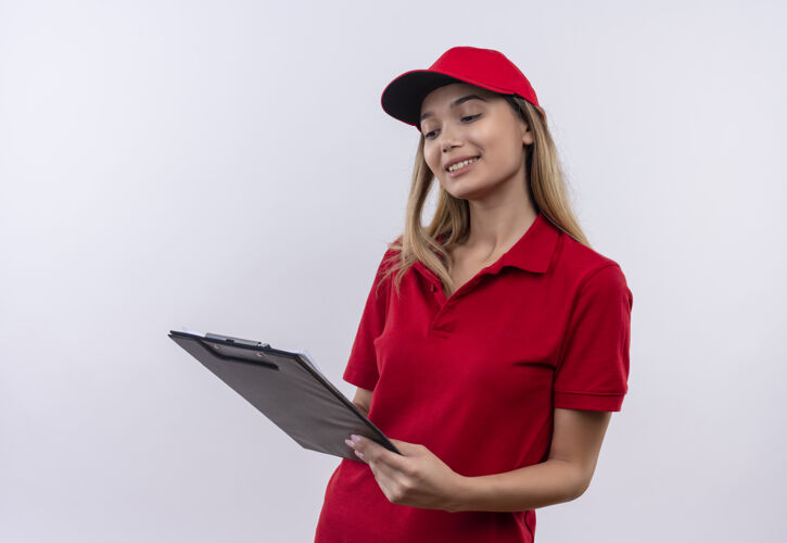 年轻高兴的年轻送货女孩穿着红色制服 戴着帽子 看着白色墙上孤立的剪贴板帽子送货制服
