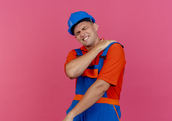 手眼睛紧闭的生病的年轻男性建筑工人穿着制服 戴着安全帽 手放在粉红色的疼痛的肩膀上安全头盔眼睛