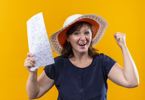 表演快乐的中年旅行家妇女 戴着帽子 拿着地图 在孤零零的黄墙上做着“是”的手势帽子地图中年