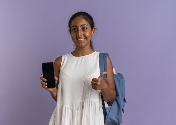 女学生带着微笑的年轻女学生 背着书包 大拇指举在紫色手机上拇指年轻紫色