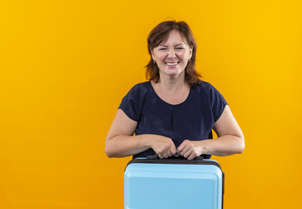 手势微笑的中年旅行家女子拿着手提箱上孤零零的黄色表情手提箱女人