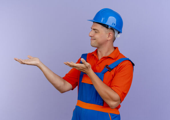 请站在侧视图高兴的年轻男性建设者穿着制服和安全帽假装持有紫色的东西建筑工安全站立
