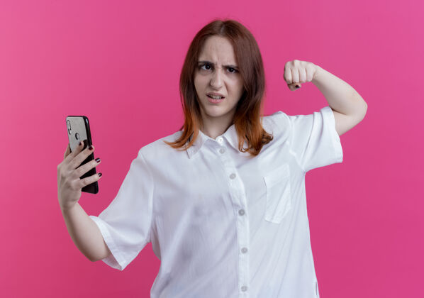 手势自信的年轻红发女孩拿着手机 表现出强烈的姿态孤立在粉红色的墙上粉红墙秀