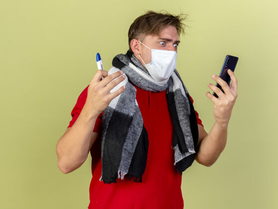 愤怒愤怒的年轻英俊的金发病男子戴着面具和围巾拿着温度计和手机看着电话隔离在橄榄绿的墙上面具体温计脸