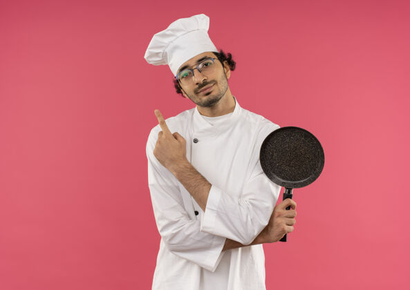 平底锅高兴的年轻男厨师穿着厨师制服 戴着眼镜拿着煎锅 指着粉红色的一面年轻空间眼镜