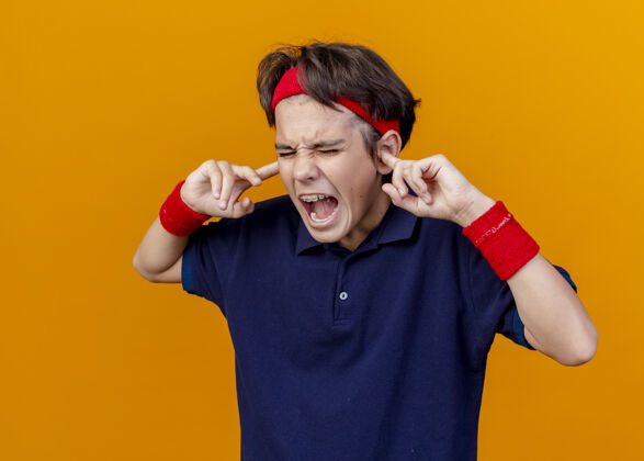 感觉愤怒的年轻英俊的运动男孩戴着头带和带牙套的腕带把手指放在耳朵里闭着眼睛孤立在橙色的墙上牙套眼睛站着