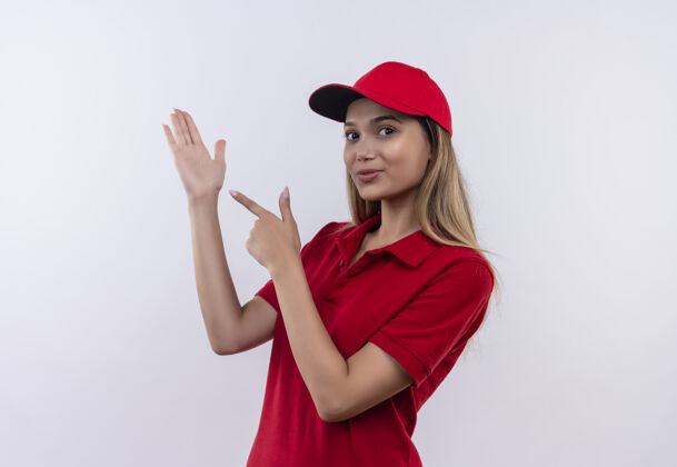请身穿红色制服 头戴鸭舌帽的年轻送货员指着隔离在白墙上的手穿制服送货
