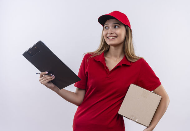 帽子看着身边穿着红色制服 戴着帽子 拿着剪贴板和盒子的年轻送货女孩 隔离在白色的墙上穿剪贴板壁板