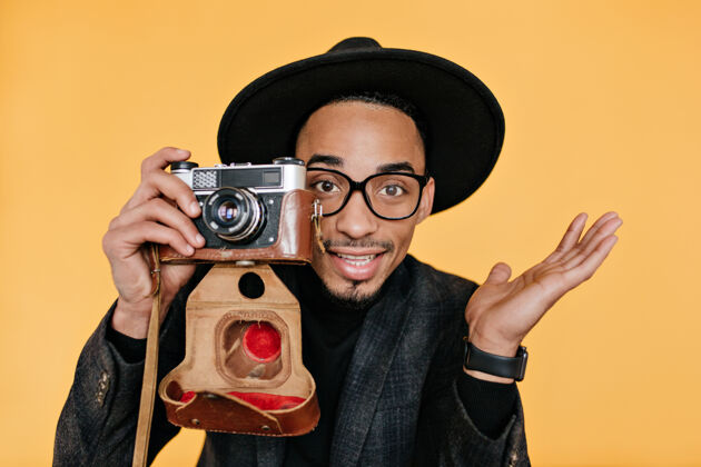 时尚灵感来源于棕色眼睛的混血儿男子拿着相机站在黄色的墙上非洲男摄影师享受工作的室内特写照片黑发年轻人快乐