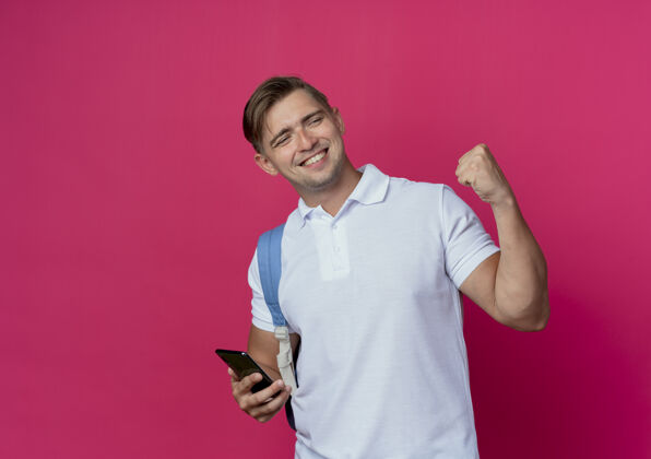 姿态看着面带微笑的年轻英俊的男学生 背着书包 拿着电话 在粉红色的墙上显示着“是”的手势壁板帅气穿