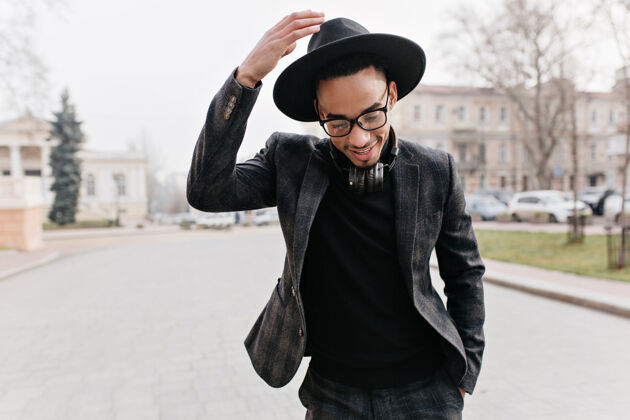 散步美丽的非洲男人在城市街道上摆出羞涩的微笑戴着帽子的时髦黑人戴着耳机站在路上大笑街道城市寒冷