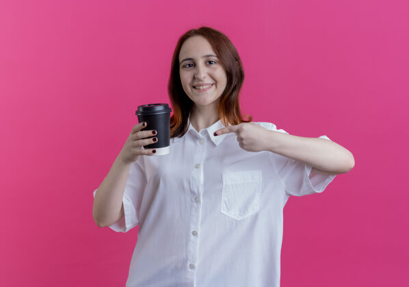 咖啡微笑着的年轻红发女孩捧着咖啡杯指着孤立的粉红色杯子女孩粉色