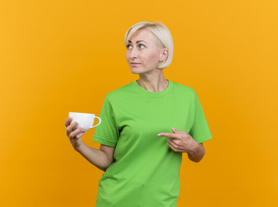 拿着自信的中年金发女人端着一杯茶 看着隔离在黄色墙上的一边站着感觉指着