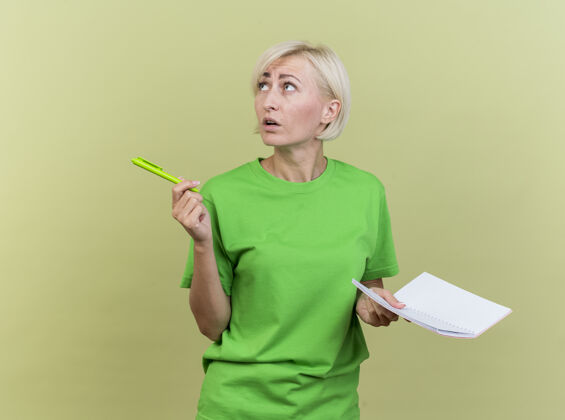 橄榄印象深刻的中年金发女人拿着笔和笔记本看着隔离在橄榄绿墙上的一面垫笔记衣服