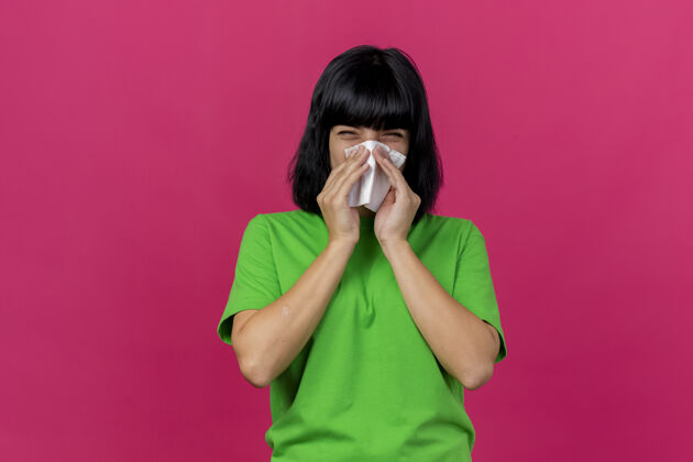 疾病愁眉不展的年轻病女人看着前面擦鼻子用餐巾隔离在粉红色的墙上餐巾皱眉人