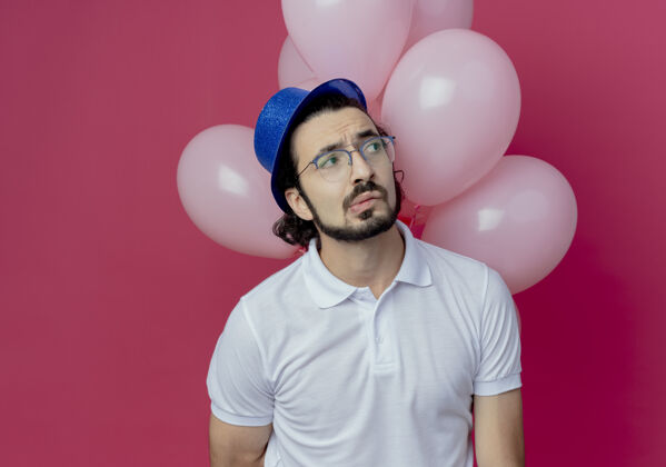 帽子看着身边迷茫的帅哥戴着眼镜和蓝色的帽子站在前面 气球孤立在粉红色的墙上困惑手势墙