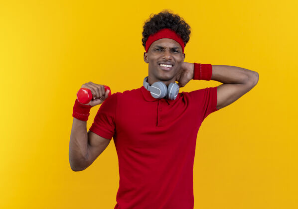 穿年轻的美国黑人运动男子戴着头带和腕带 脖子上戴着耳机 手持哑铃 手放在头后 隔离在黄色的墙上男人腕带非裔美国人