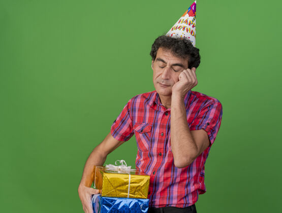 聚会无聊的中年派对男人戴着生日帽拿着礼包把手放在脸上闭着眼睛隔离在绿色的墙上抱着生日包