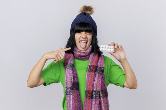 人自信的年轻生病的女人戴着冬天的帽子和围巾 展示着一包药片 指着它 看着前面 展示着隔离在白墙上的舌头姿势穿着围巾