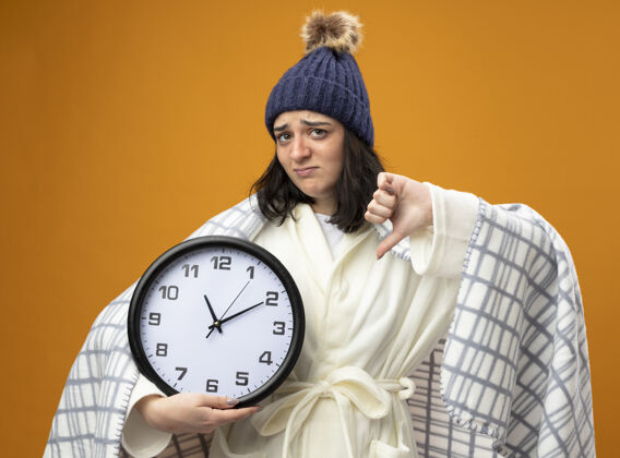 拇指年轻的生病的女人穿着长袍 戴着冬天用格子布包起来的帽子 拿着时钟看着前面 拇指朝下 孤立地站在橙色的墙上人抱穿着