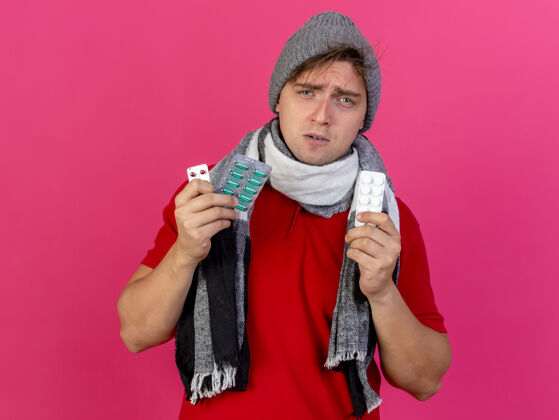 衣服愁眉不展的年轻英俊的金发病男子戴着冬天的帽子和围巾 手里拿着一包包的药丸 看着隔离在粉红色墙上的前面皱眉人市民