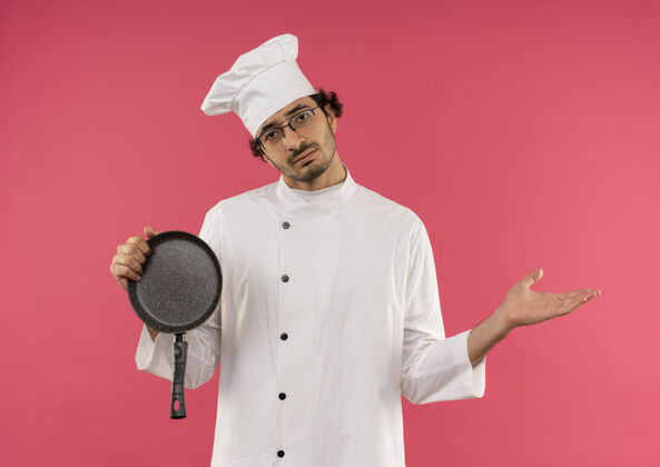 粉色悲伤的年轻男厨师穿着厨师制服 戴着眼镜 手里拿着煎锅 用手指着粉红色的一面悲伤平底锅手