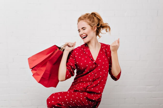 睡衣穿着红色睡衣的热情女孩手舞足蹈 纸袋孤立在白墙上滑稽的金发女人拿着新年礼物 站在砖墙附近睡眠年轻室内