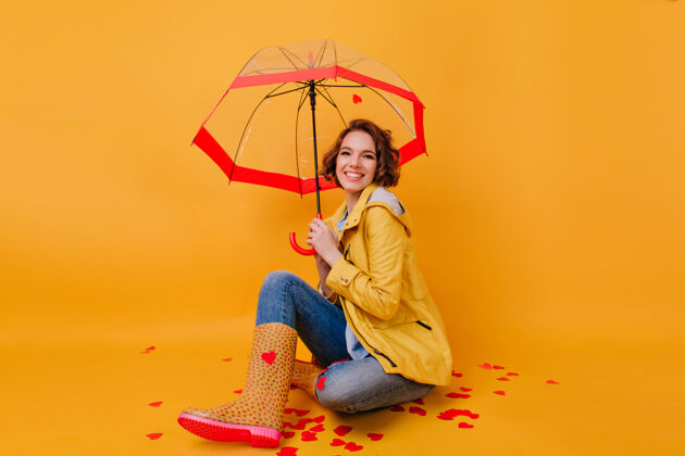 魅力穿着时髦秋装的无忧无虑的女孩在阳伞下交叉着双腿摆姿势黄色墙上挂着一幅漂亮女士打着伞笑的室内画像年轻阳伞卷发