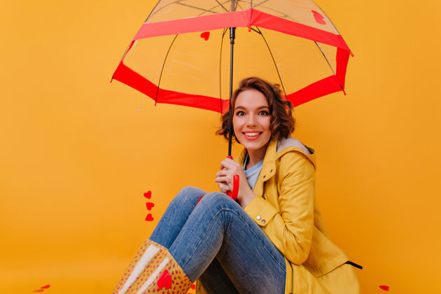 阳伞穿着胶鞋的迷人卷发女孩在雨伞下愉快地摆着姿势黄色墙壁上隔离着微笑的可爱秋装女人的室内肖像室内肖像优雅