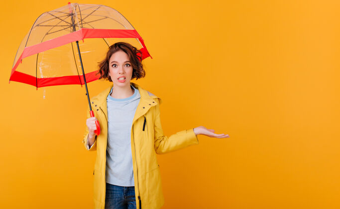 阳伞黄色墙壁上穿着雨衣拿着雨伞的时髦女孩的室内肖像卷曲的年轻女士拿着阳伞表示惊讶优雅表情成人