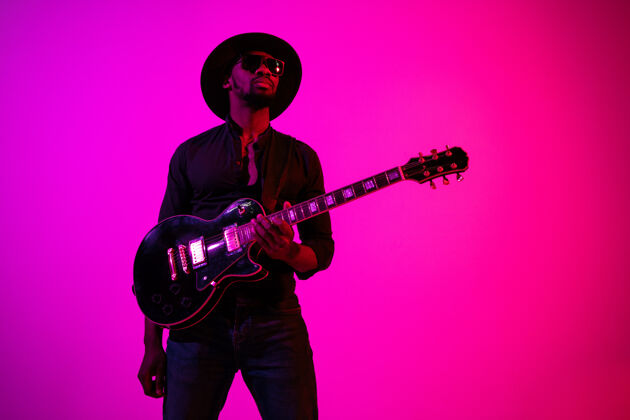 吉他手年轻的非洲裔美国音乐家在霓虹灯下像摇滚明星一样在渐变的紫粉色背景上弹吉他男士衬衫黑色