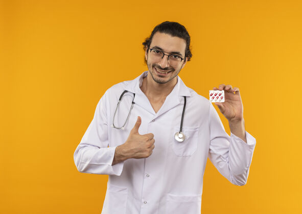 向上微笑着的年轻医生戴着医用眼镜穿着医用长袍手持听诊器拿着药片他的大拇指朝上放在黄色拇指医学医生