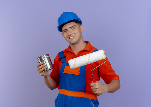 年轻面带微笑的年轻男性建筑工人身穿制服 头戴安全帽 手持油漆罐和油漆辊上的紫色油漆油漆安全头盔