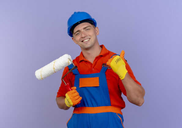 建筑工人面带微笑的年轻男性建筑工人身穿制服 戴着安全帽 戴着手套 拿着油漆辊 大拇指朝上放在紫色上油漆紫色微笑