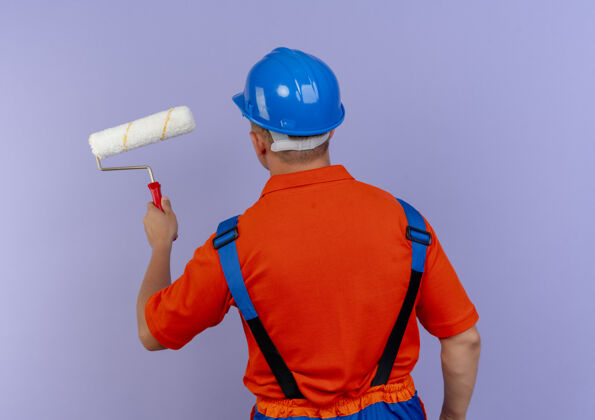 站立站在后面看年轻的男建筑工人穿着制服 戴着安全帽 手里拿着紫色的油漆辊紫色年轻油漆
