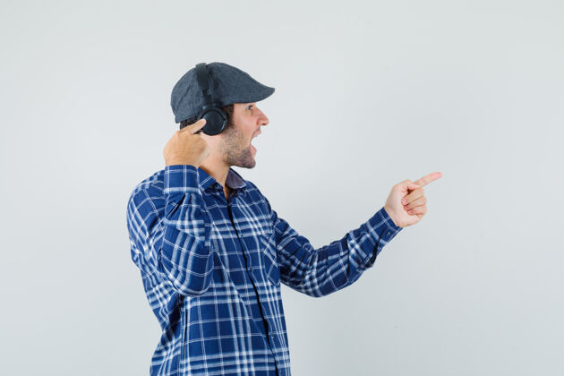 衬衫年轻人指着他的耳机 站在一边 穿着衬衫 戴着帽子 看上去很快乐 前面的景色朋友耳机成功