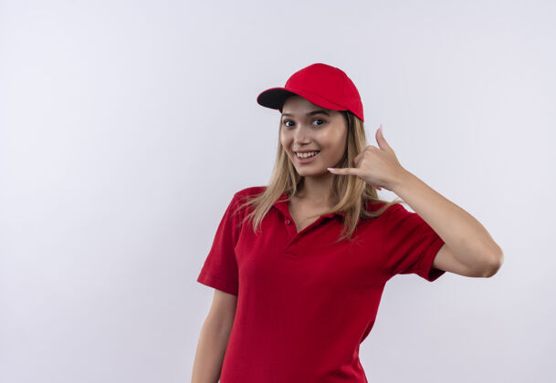 电话微笑着的年轻送货女孩穿着红色制服 戴着帽子 在白色衣服上显示着电话的手势年轻表情制服