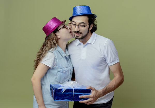 持有一对年轻夫妇戴着粉色和蓝色的帽子 女孩在男孩的耳朵上低语 男孩拿着礼盒孤立在橄榄绿上穿帽子粉色