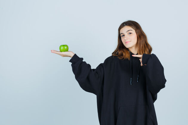 壁板年轻女士穿着超大的连帽衫 裤子拿着苹果 同时指着左边 看起来很高兴 正面视图苹果而指尖